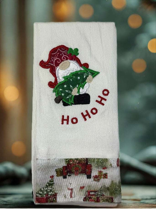 White cotton flour sack towel with embroidered gnome holding Christmas tree and red "Ho Ho Ho" text with holiday print hem
